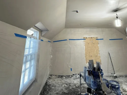 Dormer room mid-job, walls masked in plastic and a mixing station set up under a fresh plaster ceiling