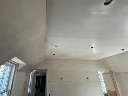 Plastered room with sloped ceilings and two dormer windows, plywood left at the base for wainscoting