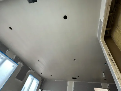 Plastered ceiling above walls still in blueboard, windows masked and a crew member at work below