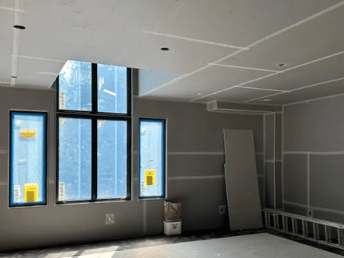 Great room hung in blueboard with taped joints on walls and ceiling, windows masked before plastering