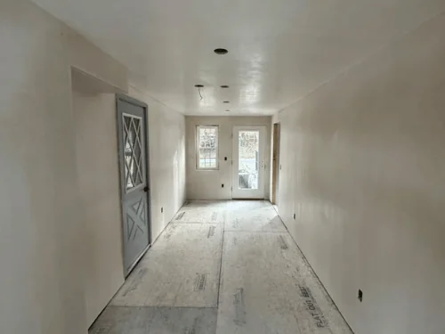 Long room with smooth plaster walls running to a pair of glass doors, floor covered with protection board