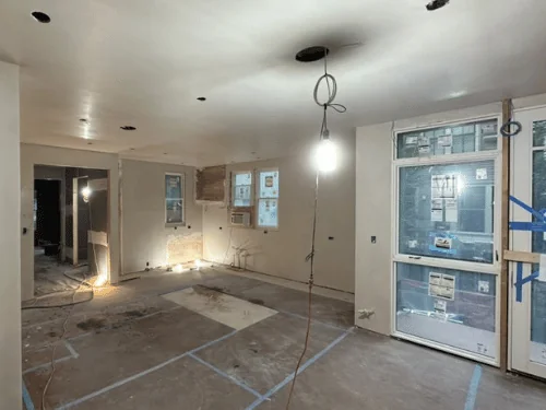 Living area with smooth plaster walls and ceiling, new windows still labeled and the floor covered