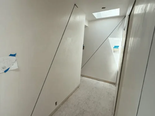 Hallway wall in white plaster crossed by angled reveal lines, a skylight at the far end