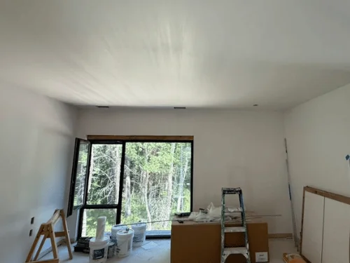 White plaster room with a large corner window to the woods, ladders and buckets still on site