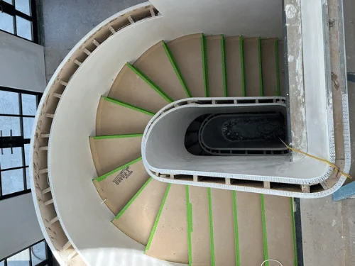 Curved plaster stair walls wrapping an oval stairwell, treads protected with board and tape