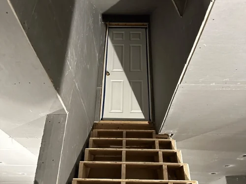 Stairwell sheathed in blueboard with taped joints, leading down to a paneled door