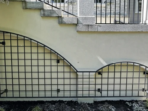 Smooth cream stucco wall under a granite stair, fitted with a black arched trellis
